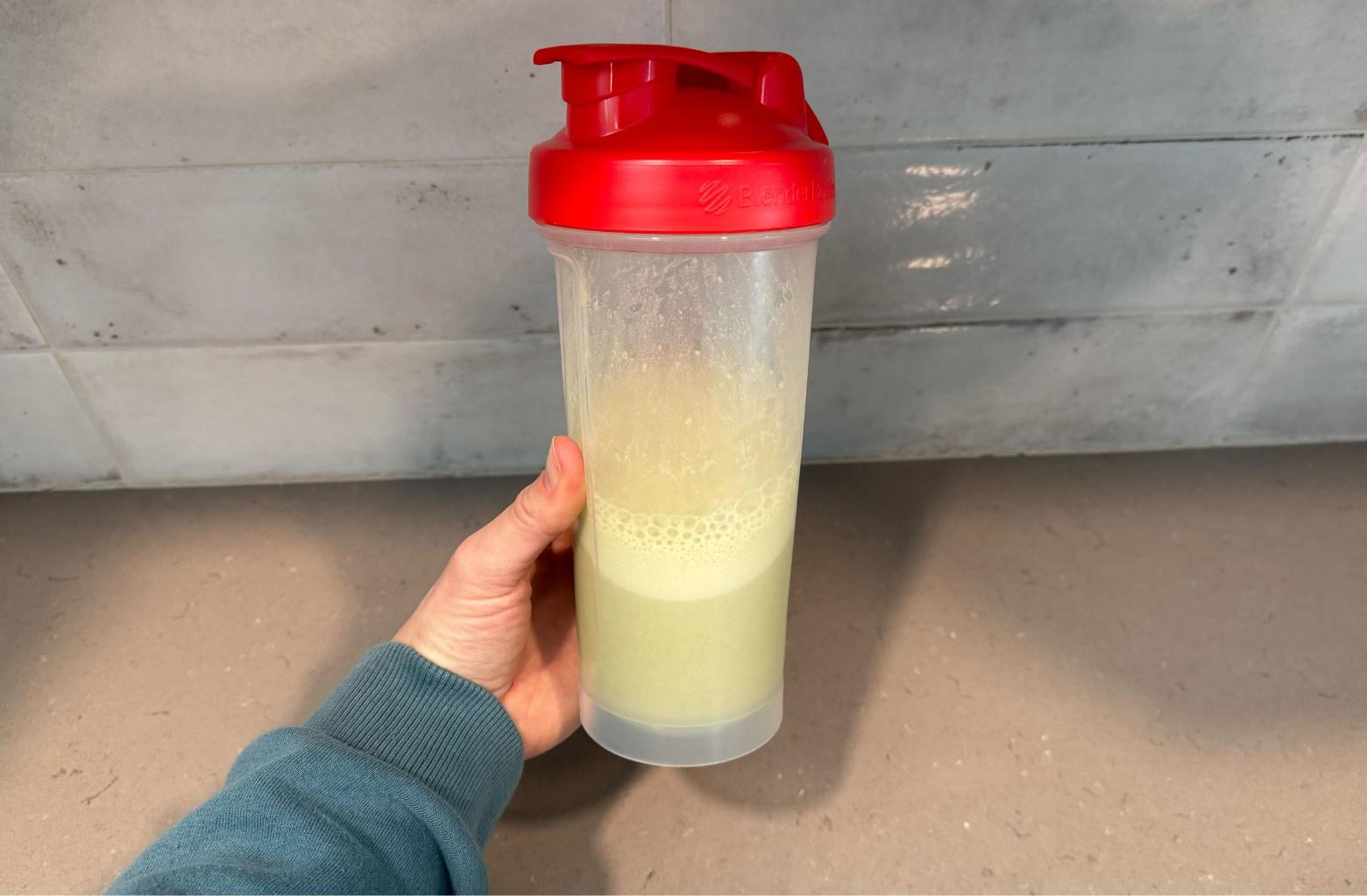 A person holds a shaker bottle filled with vanilla Vega Protein & Greens powder mixed with cold water.